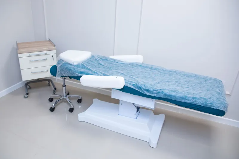 A white medical exam room with a padded exam table and a rolling cabinet