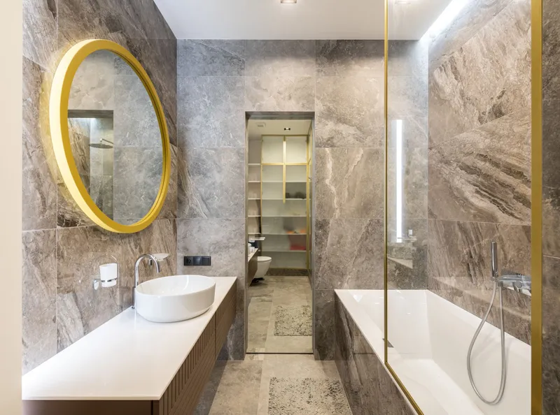 A bathroom with stone-look tile, a round gold mirror, a vessel sink and a glass-screened tub