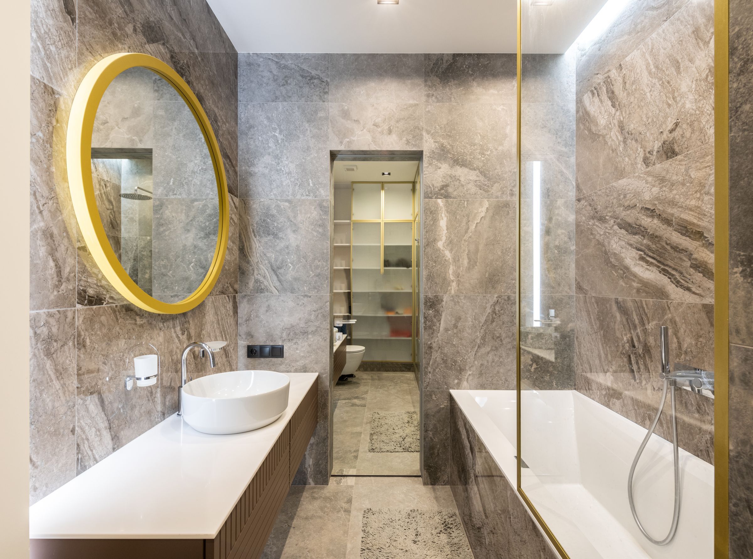 A bathroom with stone-look tile, a round gold mirror, a vessel sink and a glass-screened tub