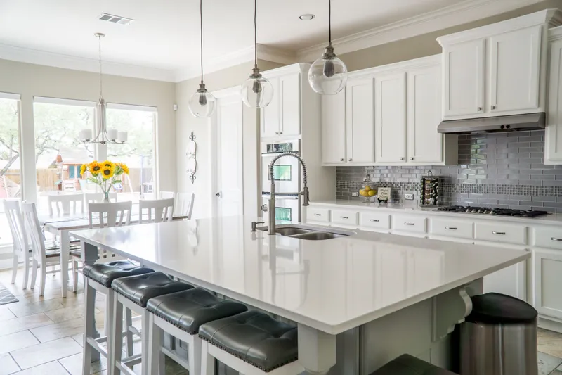 A bright white kitchen with a long quartz island, pendant lights and a dining table by the windows