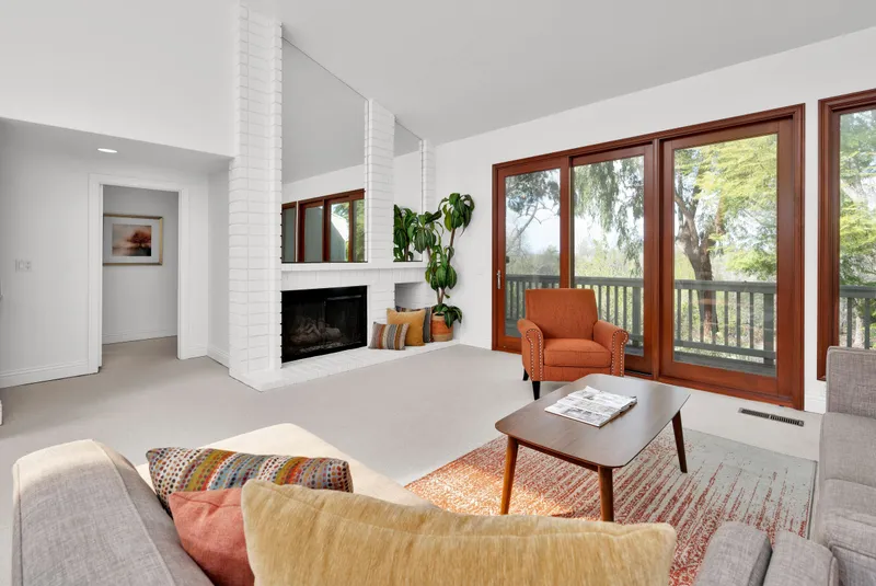 A bright living room with a white brick fireplace, an orange armchair and tall wood-framed windows