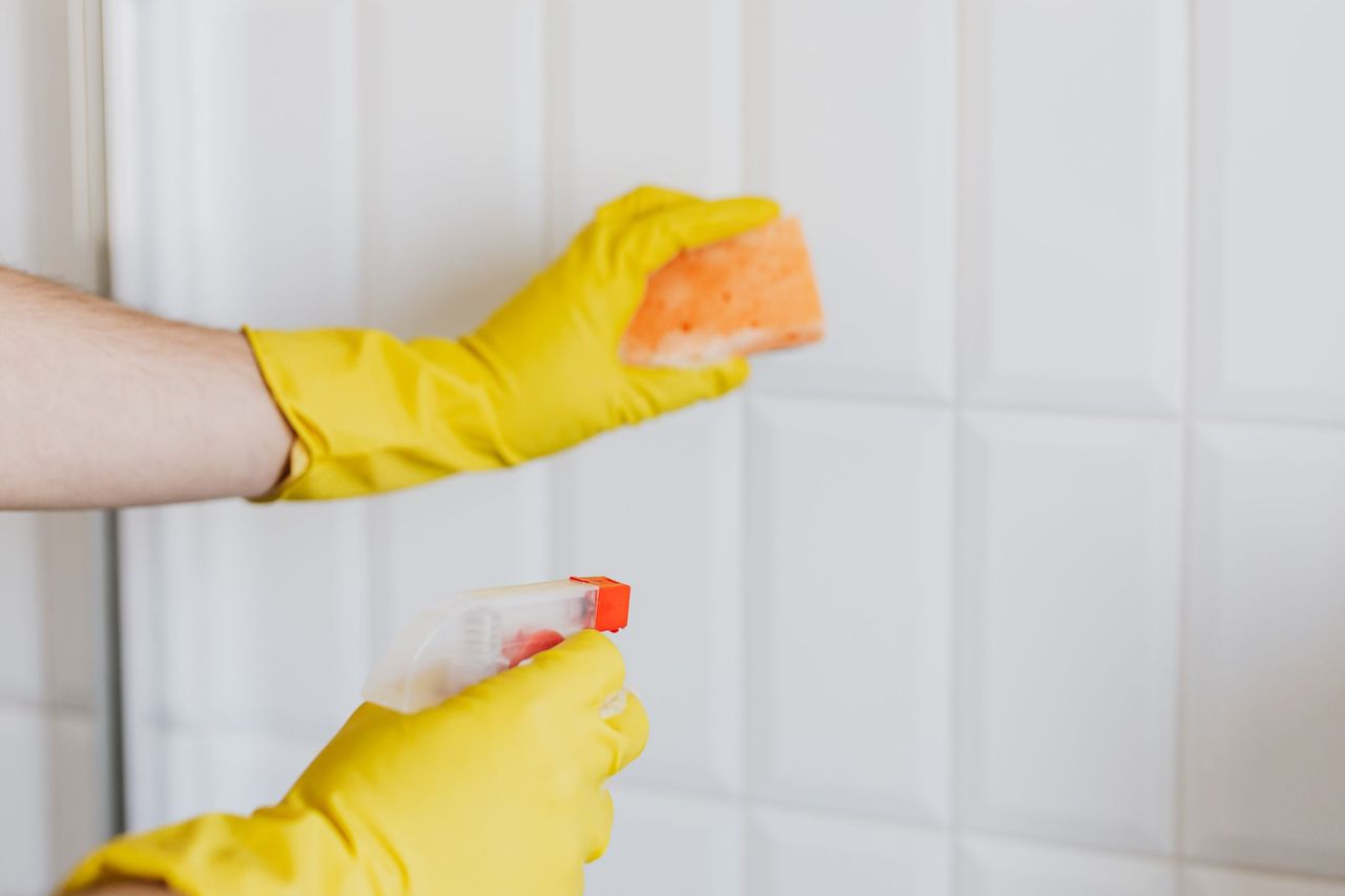 Yellow-gloved hands scrubbing white wall tile with a sponge and a spray bottle