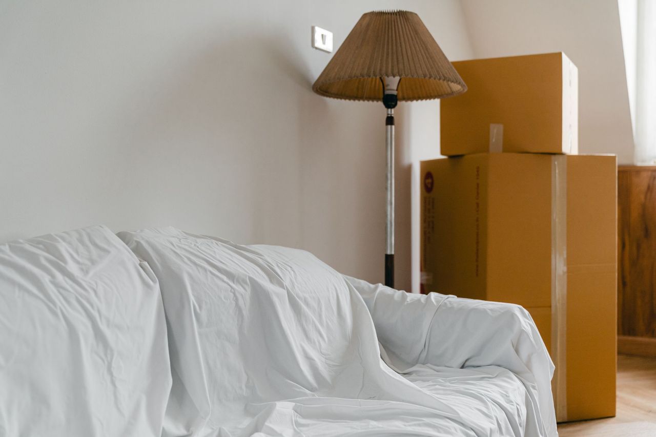 Moving boxes, a floor lamp and a sheet-covered sofa in a bright, empty room