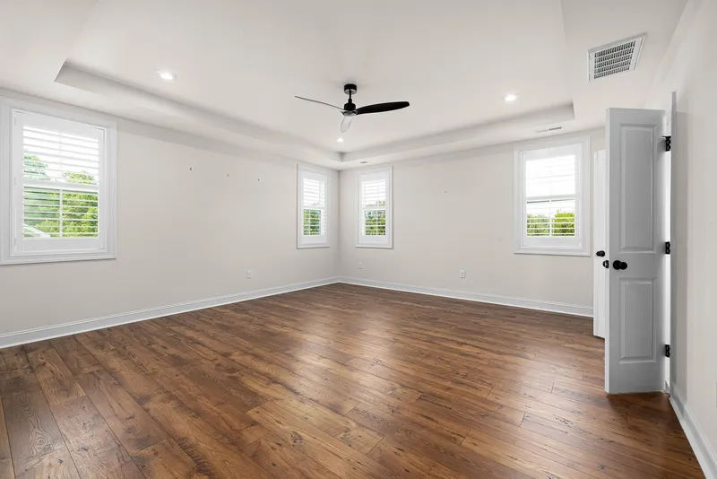 An empty bedroom with wood floors, a ceiling fan and three sunlit windows