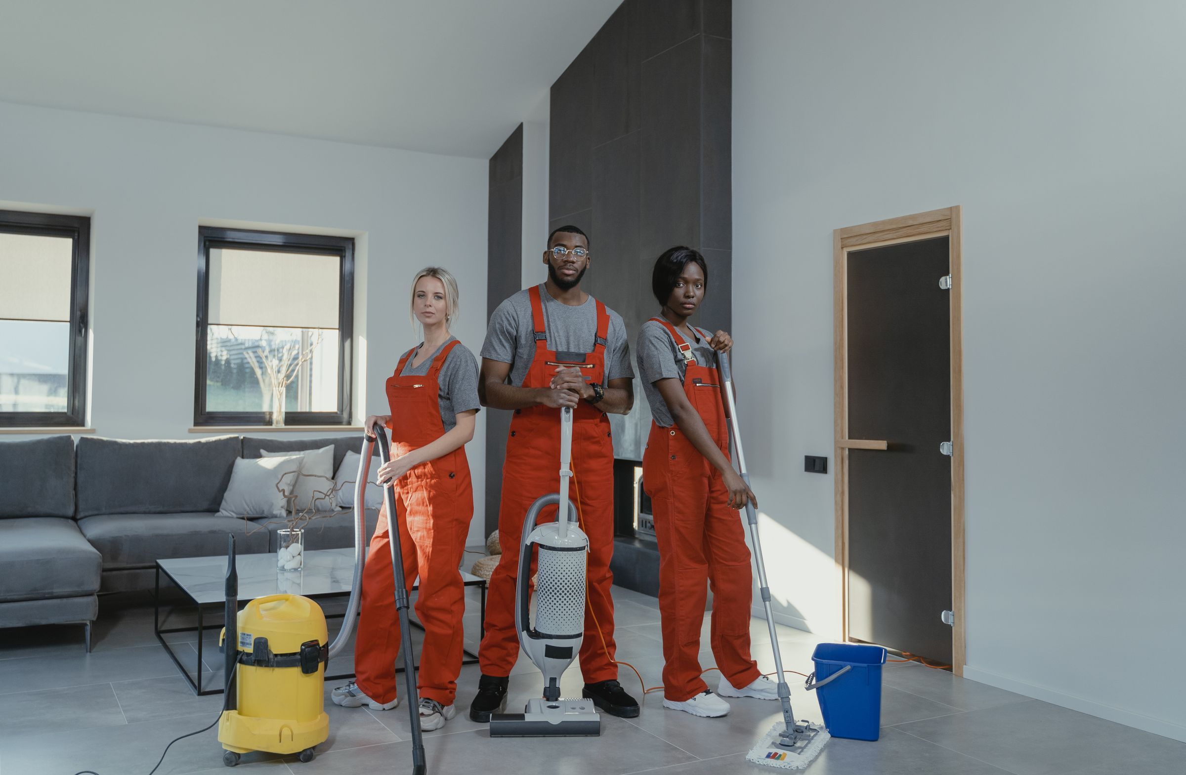 Three cleaners in orange overalls standing in a living room with a vacuum, a wet vacuum and a mop
